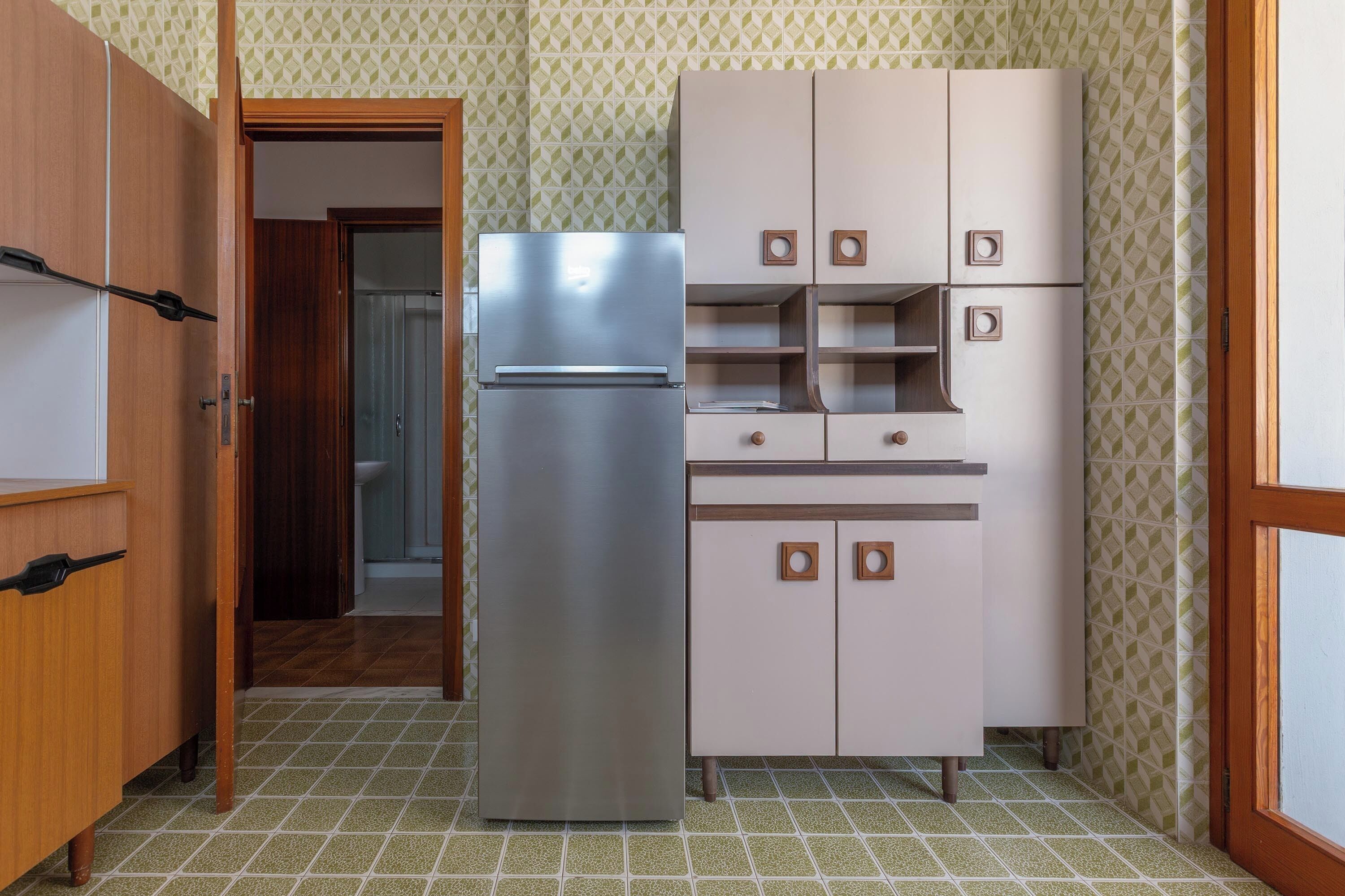 Eine kleine Küche mit Edelstahlkühlschrank, Holzschränken und Wandpapier mit Muster.
