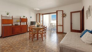 A room with a dining table, chairs, a sofa, a TV, a wooden cabinet, and a view of the outdoors. - Balconcino Vista Mare by BarbarHouse (San Foca)
