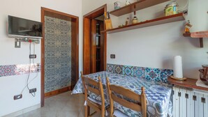 Een slaapkamer met een bed, houten stoelen, een aan de muur bevestigde televisie, planken met keukengerei en een kamerkleed met patroon.