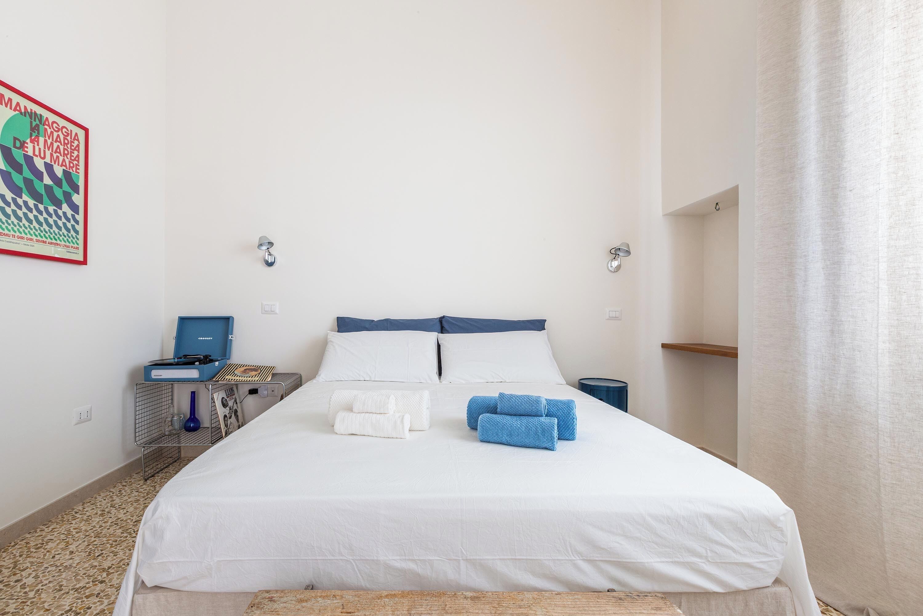 Un letto rifatto con lenzuola bianche e azzurre, un comodino con una lampada e una mensola a muro.