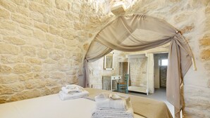 A room with a stone wall, a bed with a beige canopy, a chair, and a bathroom visible through an open door. - Trullo Pumo by Perle di Puglia (San Michele Salentino)