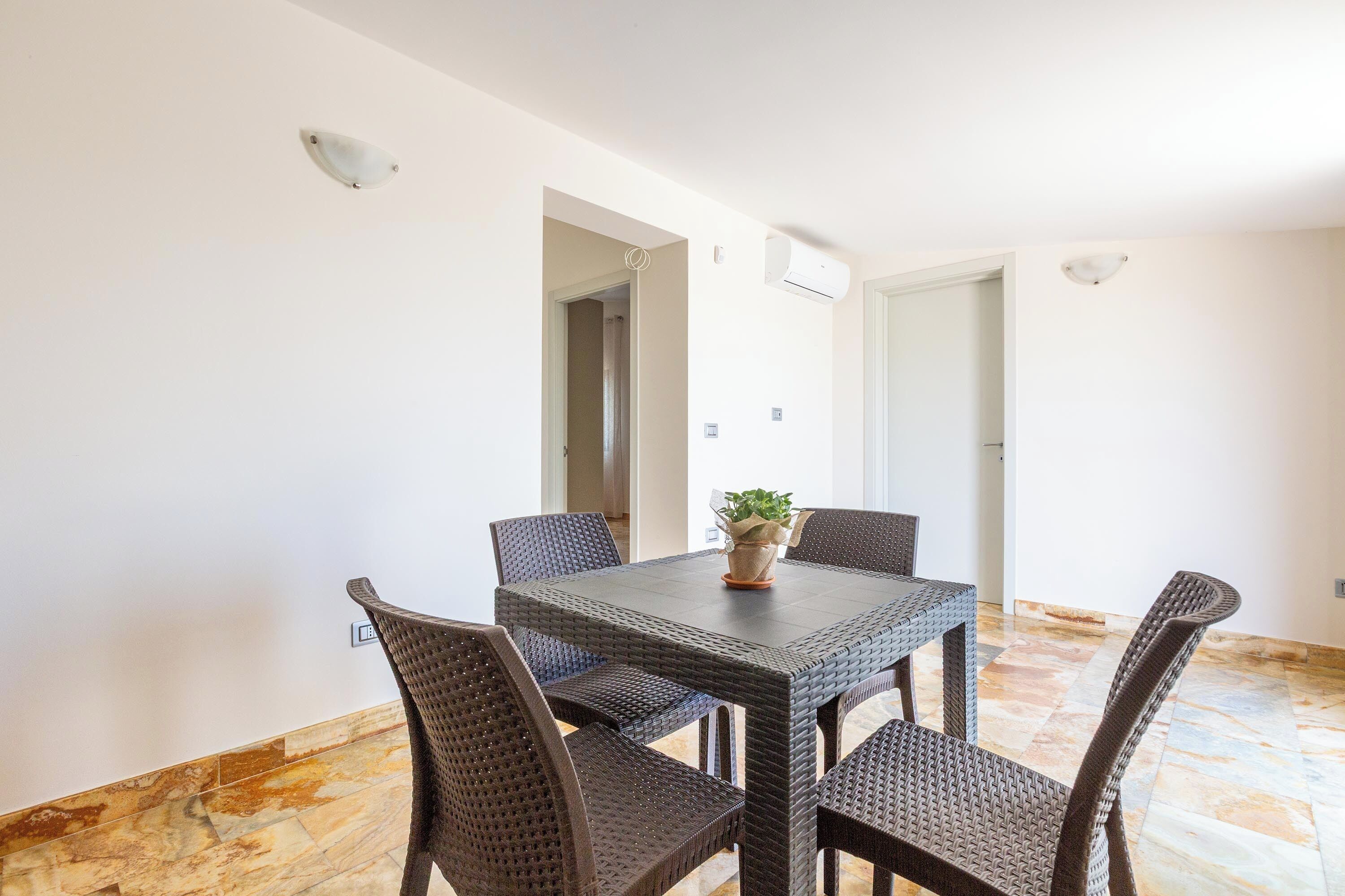 Un coin repas avec une table carrée et six chaises en osier, une plante en pot posée sur la table et un sol carrelé.