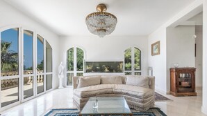 Un salón amplio con un gran ventanal, un sofá blanco, una mesa de centro de vidrio y una araña.