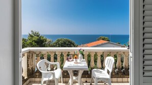 Un balcón con una mesa y sillas blancas, con vistas a un paisaje marino y zonas verdes.