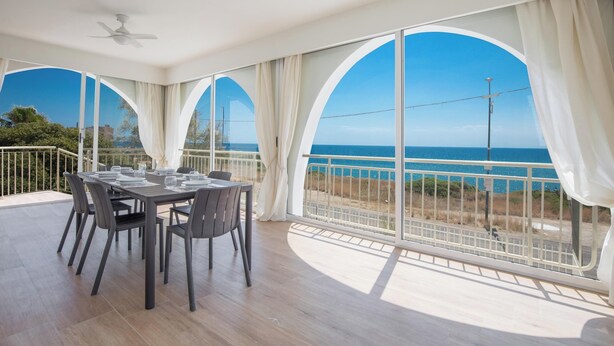 Un balcon spacieux avec une table dressée pour les repas, de grandes fenêtres en arc de cercle offrant une vue sur la mer et un ventilateur de plafond.