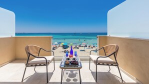 Een balkon met twee rieten stoelen, een tafel met eten en drinken, en uitzicht op een strand met parasols en mensen.