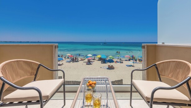 Een balkon met twee rieten stoelen en een tafel met een fruitschaal en glazen. Het uitzicht toont een strand met parasols en mensen.