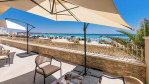 Una terraza con asientos al aire libre, un gran parasol y vista a una playa con sombrillas y gente.