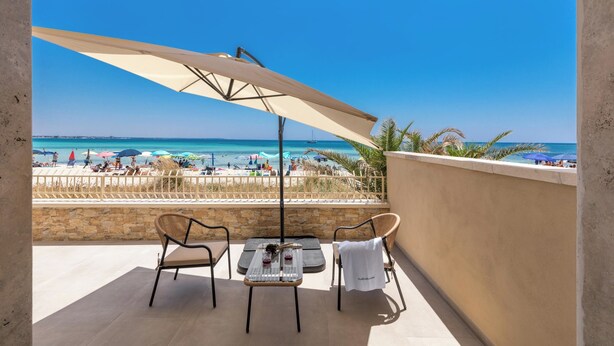 Un balcón con mesa y sillas, con vista a una playa con sombrillas y gente.