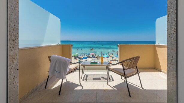 Een balkon met twee stoelen en een tafel, uitkijkend op een strand met mensen en parasols.