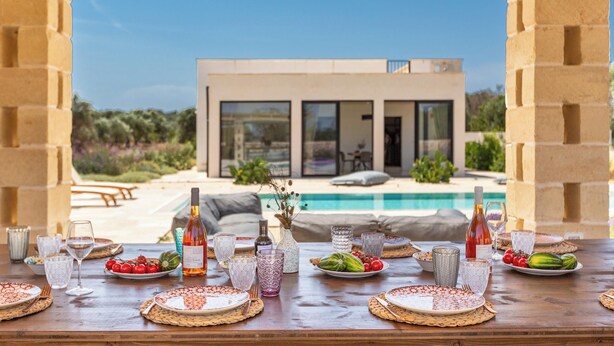 A dining table set for a meal with a view of a modern house and a swimming pool. - Villa Astra by Perle di Puglia (Ostuni)