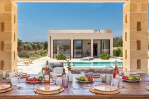 A dining table set for a meal with a view of a modern house and a swimming pool. - Villa Astra by Perle di Puglia (Ostuni)