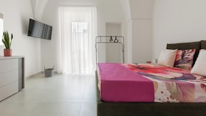 Ein Schlafzimmer mit Bett, Wandfernseher, Pflanze und einem Fenster mit Vorhängen.