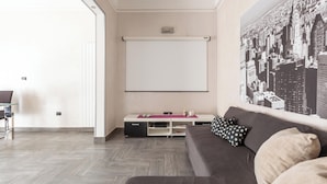Ein modernes Wohnzimmer mit einem großen Flachbildfernseher an der Wand, einer Couch mit Kissen, einem Couchtisch und einem Stadtbild als Wandkunst.
