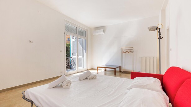 Een slaapkamer met een bed, een rode stoel, een klein tafeltje en een raam met jaloezieën.