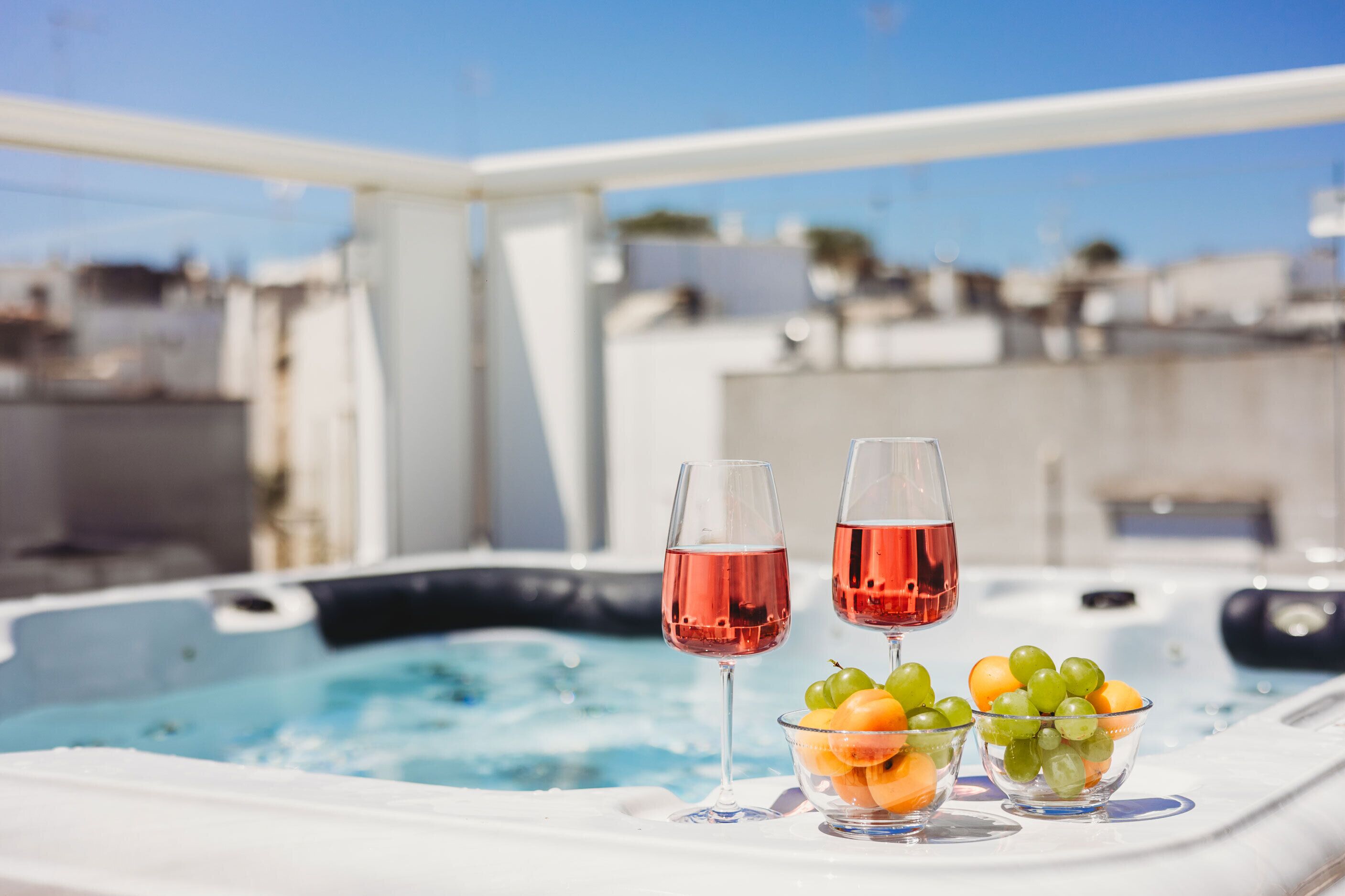 A hot tub with two glasses of red wine and bowls of fruit on a white table.