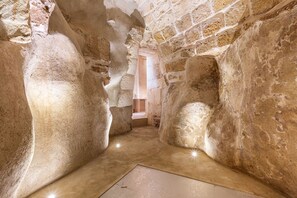 A narrow passageway with rough stone walls and a tiled floor, leading to a brighter area with a door. - Palazzo Alemar Jacuzzi & SPA by Perle di Puglia (Carovigno)