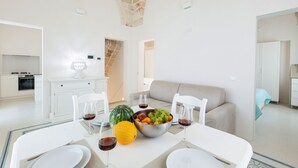 Un coin repas moderne avec une table blanche dressée pour deux personnes, un panier de fruits et deux verres de vin rouge.