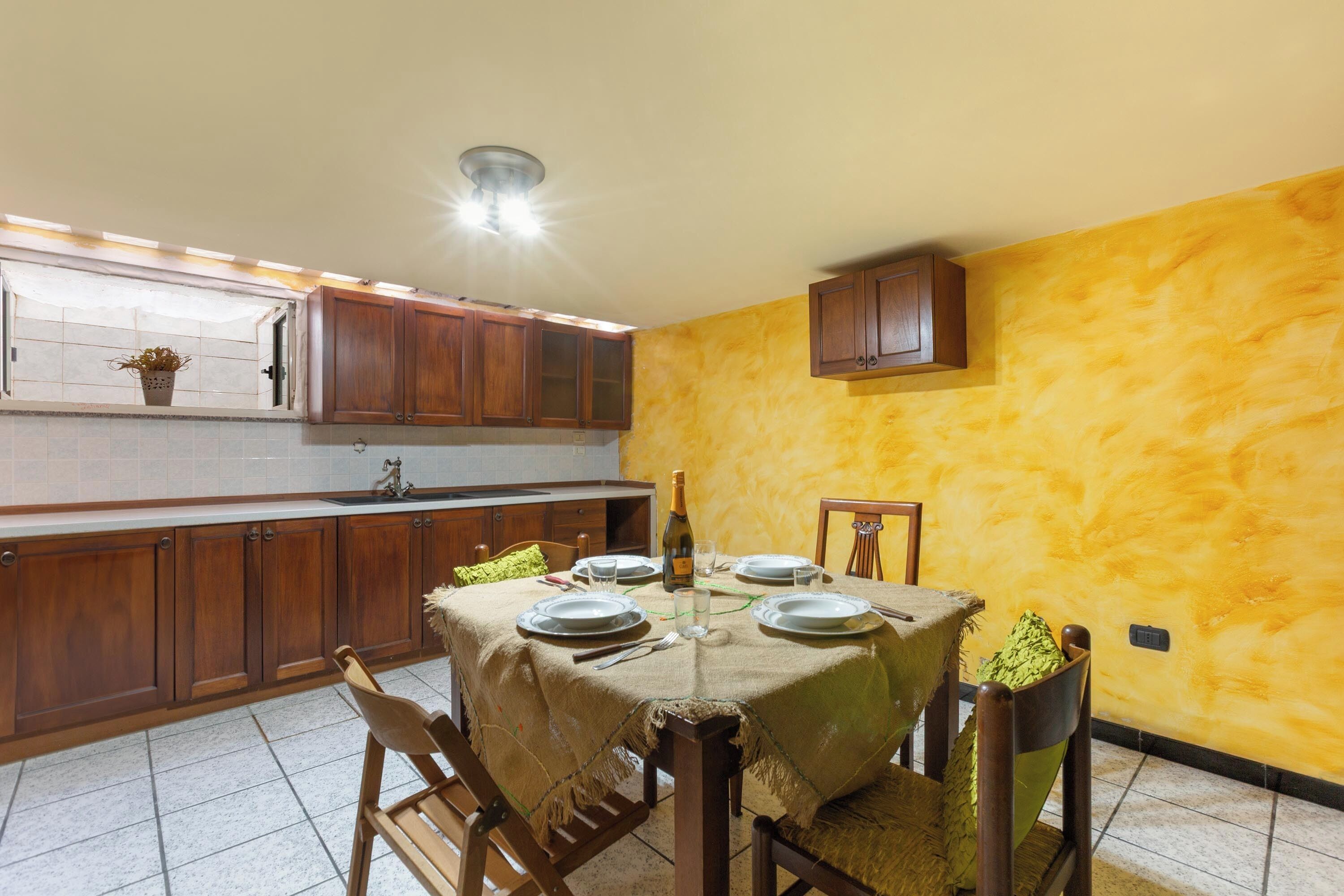 Zona pranzo con tavolo apparecchiato per quattro persone, una bottiglia di vino e cucina con mobili in legno e parete gialla.