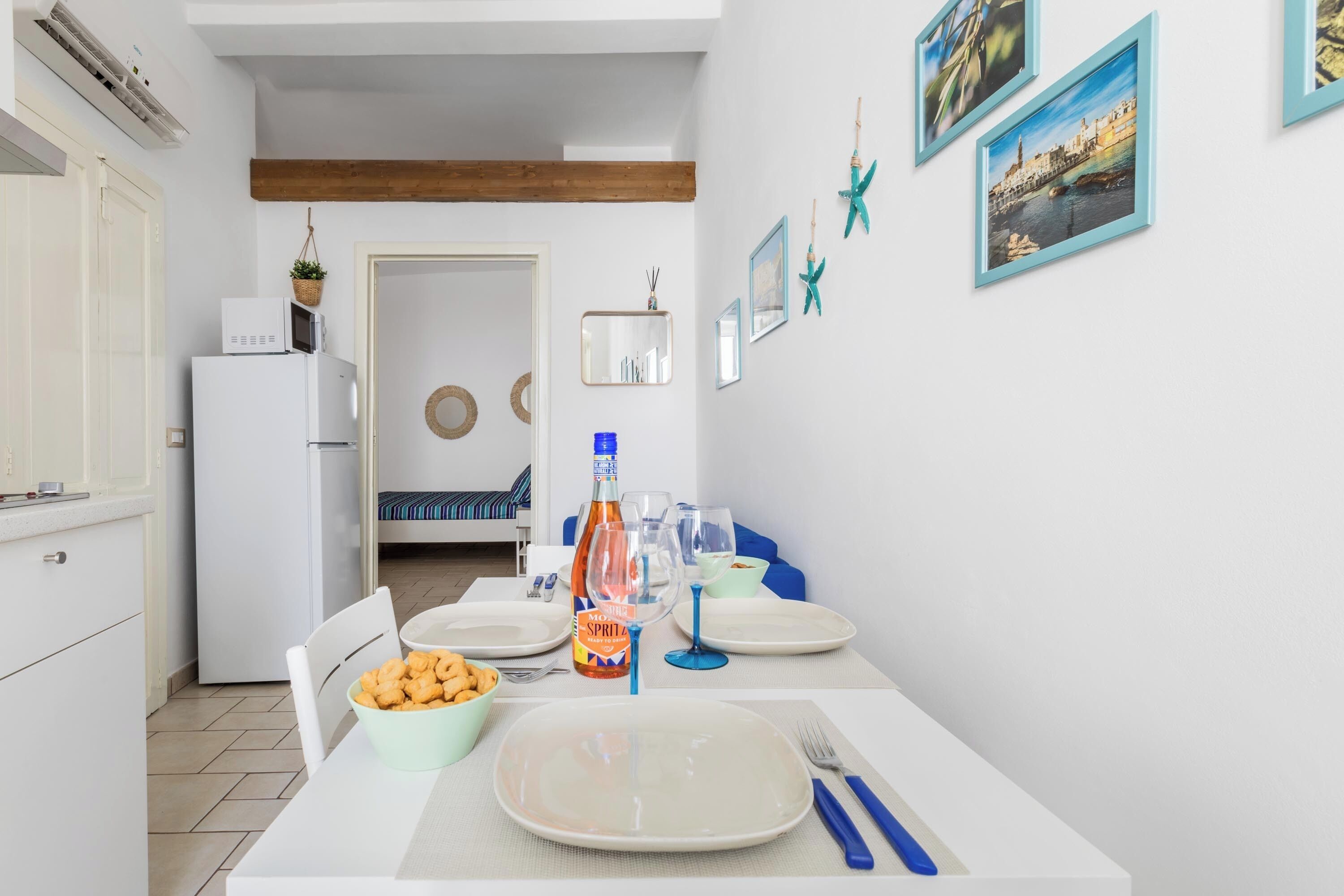 Un coin repas avec une table dressée pour deux personnes, une bouteille de Spritz et un bol de collations.