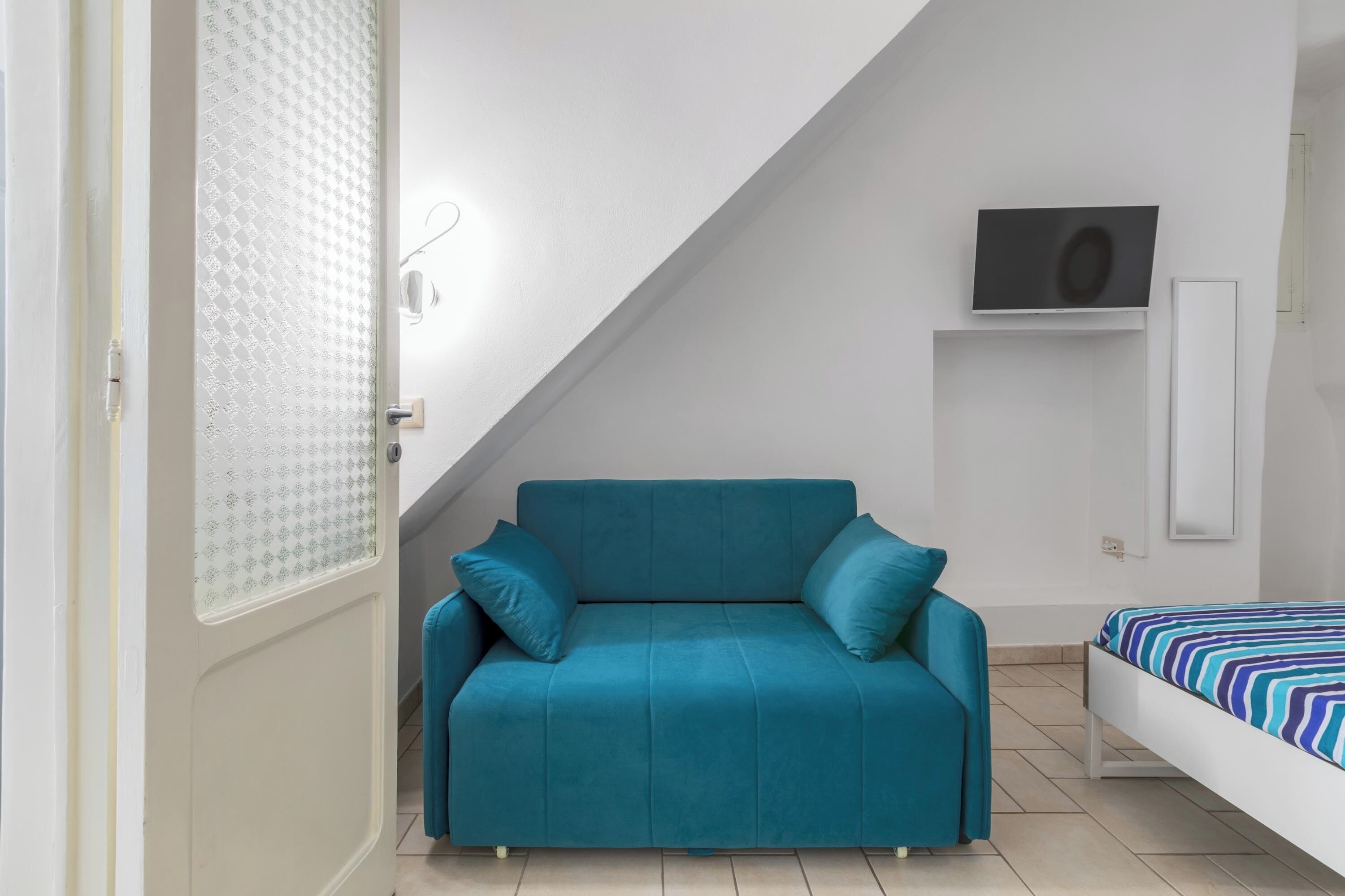 A room with a teal sofa, a bed with a striped bedspread, a TV mounted on the wall, and a patterned door.