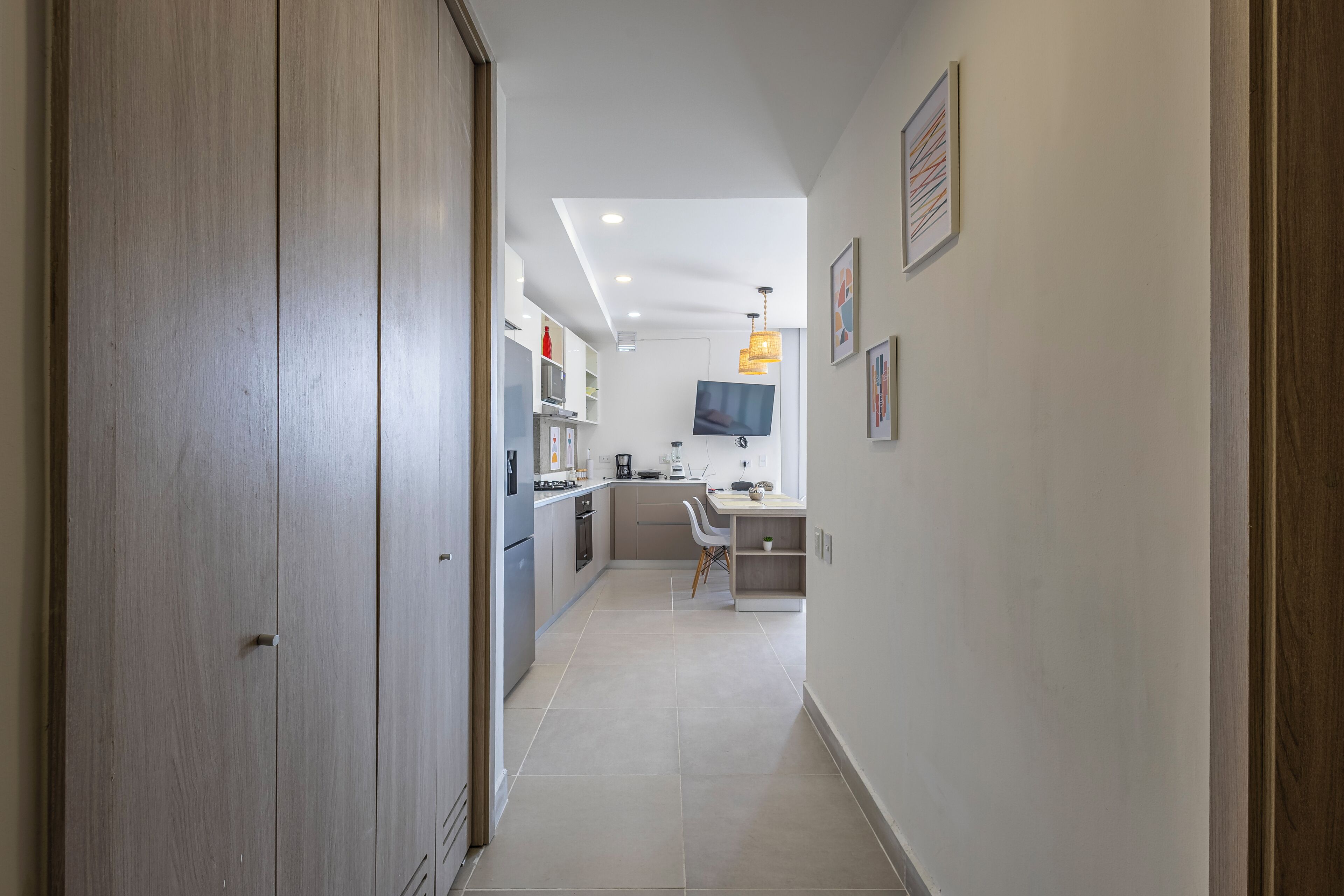 A narrow hallway leading to a kitchen with modern appliances, a dining area, and a TV mounted on the wall.