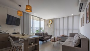 Un salon moderne comprenant un canapé, une table à manger avec des chaises, un téléviseur fixé au mur et une grande fenêtre donnant sur la ville.