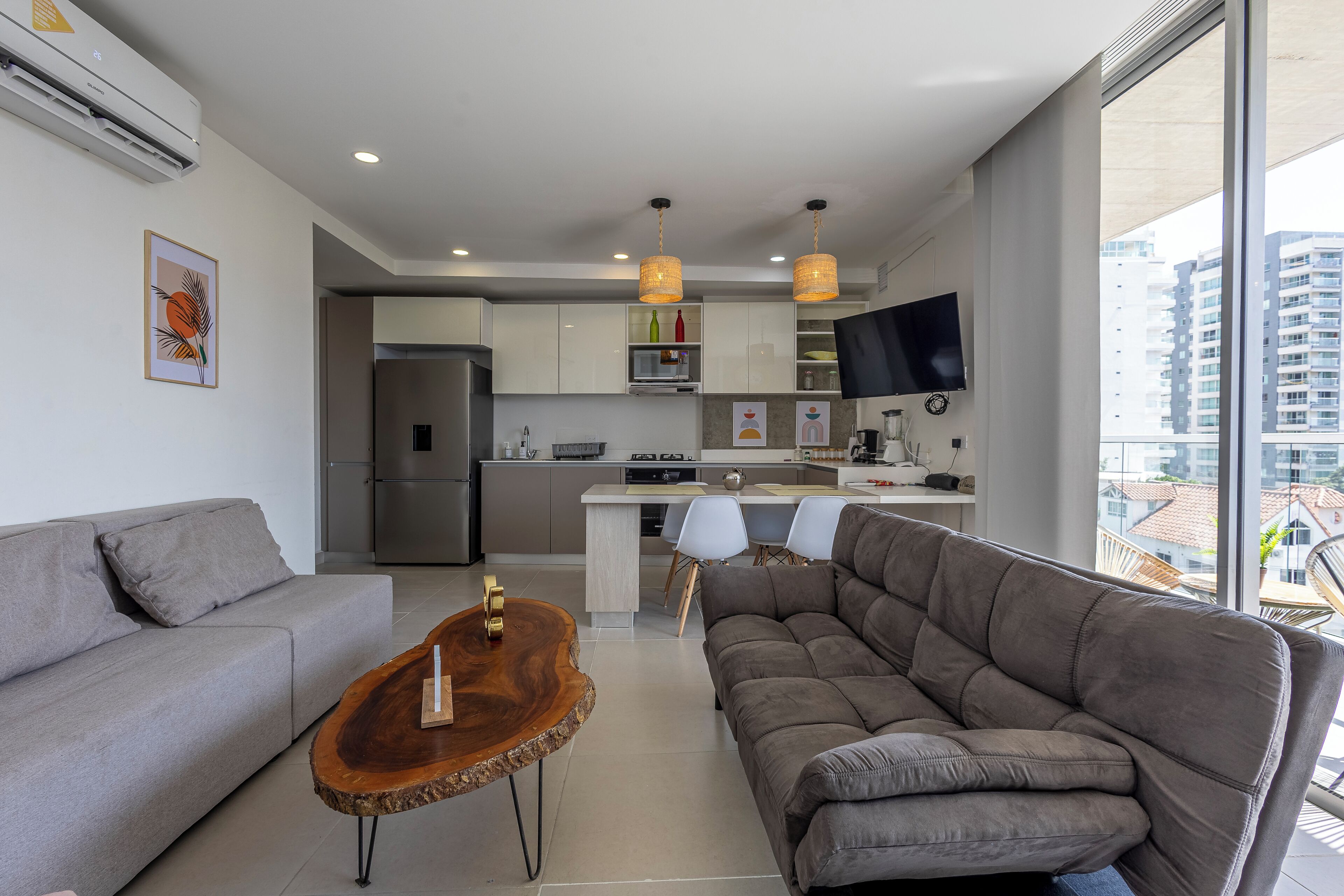 Un salon moderne avec un canapé gris, une table basse en bois et un canapé d’angle gris. On aperçoit à l’arrière-plan une cuisine avec des meubles blancs et un téléviseur à écran plat.
