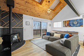 A living room with a fireplace, a sofa, a wooden ceiling, and a large window. - Camelback - Game Room, Near Hiking & Waterparks (Tannersville)