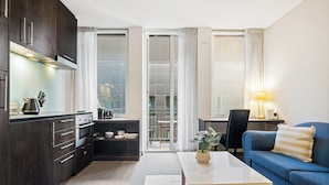 A modern kitchen with dark cabinets, a dining area with a blue sofa and a table, and a sliding glass door leading to a balcony. - Stylish Central Studio With Gym (Melbourne)