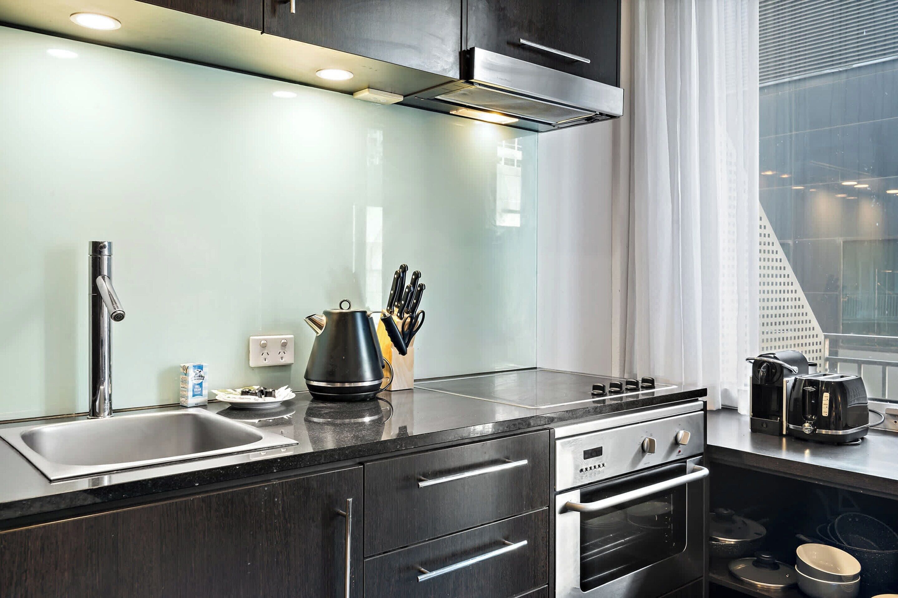A modern kitchen with a stainless steel sink, a kettle, a coffee maker, and a window with a view of a building.