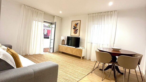 Quarto de hotel moderno com um sofá cinzento, uma mesa de jantar de madeira com quatro cadeiras, uma TV de tela plana sobre um móvel de madeira e uma janela grande com cortinas brancas.