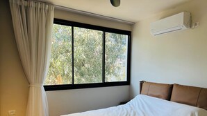 Un dormitorio con cama, ventana con vista a árboles y aire acondicionado en la pared.