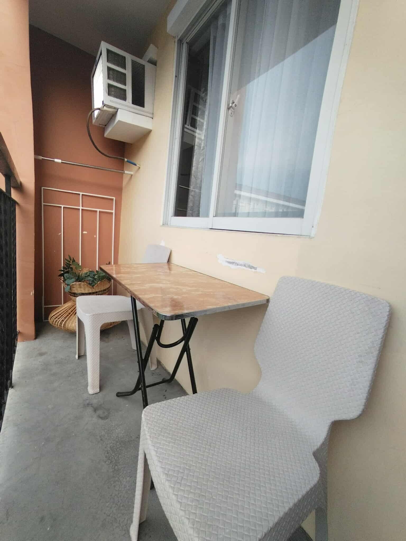 Un pequeño balcón con una mesa de madera y dos sillas blancas, un equipo de aire acondicionado y una ventana con cortina.