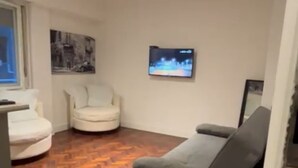 Een kamer met een grijze bank, een witte fauteuil, een televisie aan de muur en een houten vloer.