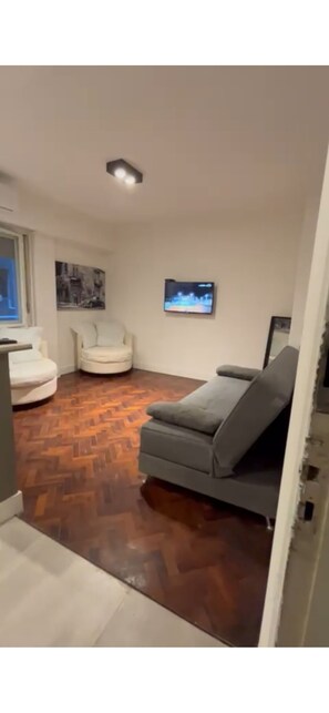 Ein Zimmer mit grauem Sofa, einem weißen Sessel, einem an der Wand befestigten Fernseher und Holzboden.