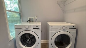 A laundry room with two white LG front-loading washing machines, a small window, and a metal shelf with clothes. - Delightful 3-bedroom house in charming Tucker (Tucker)