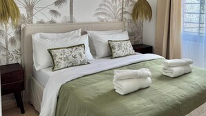 Ein ordentlich bezogenes Bett mit weißen Laken und einer grünen Tagesdecke, zwei dekorative Kissen mit Blumenmuster und ein Stapel gefalteter Handtücher darauf.