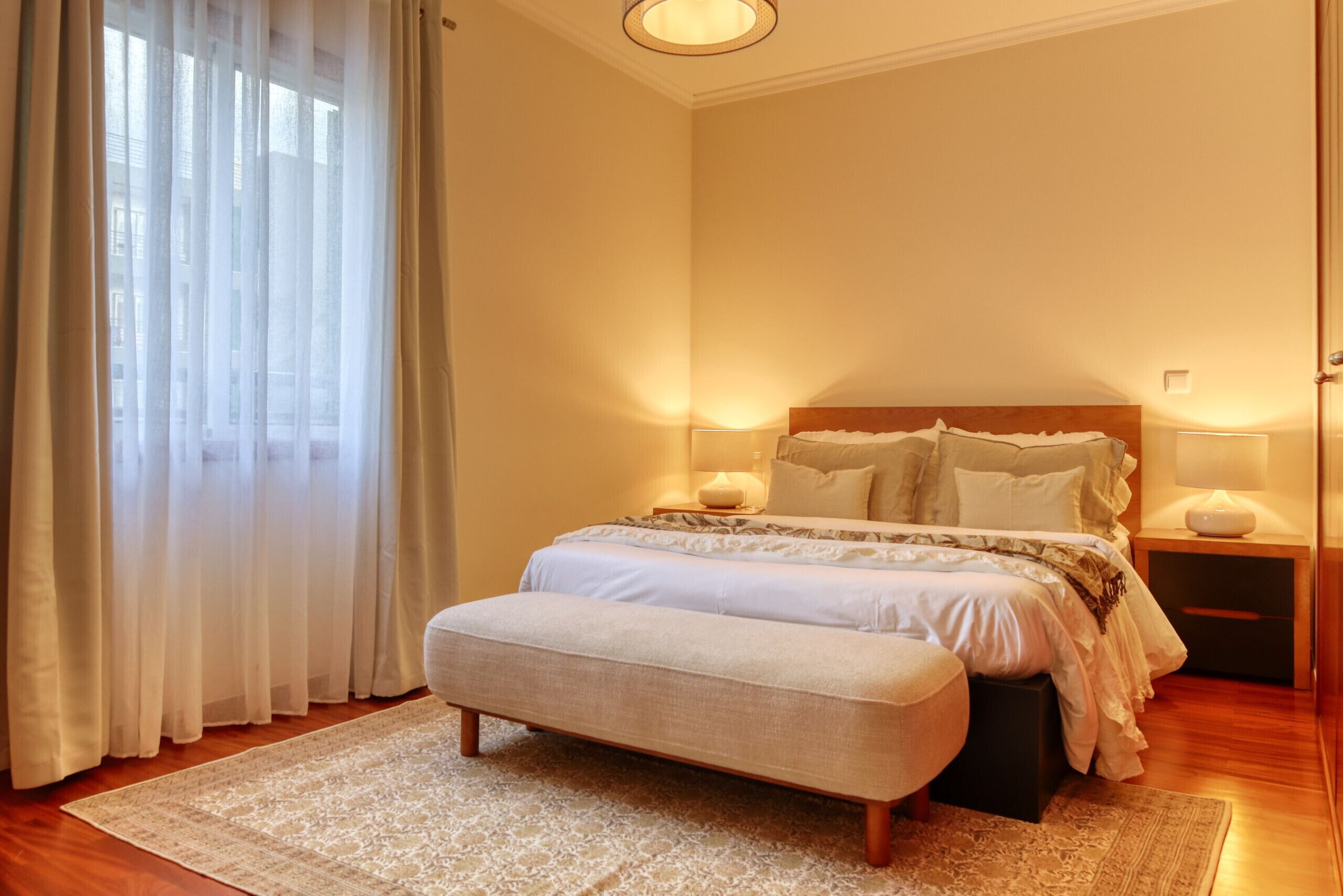 Un dormitorio con una cama grande, lámparas de noche, un banco y una ventana con cortinas translúcidas.