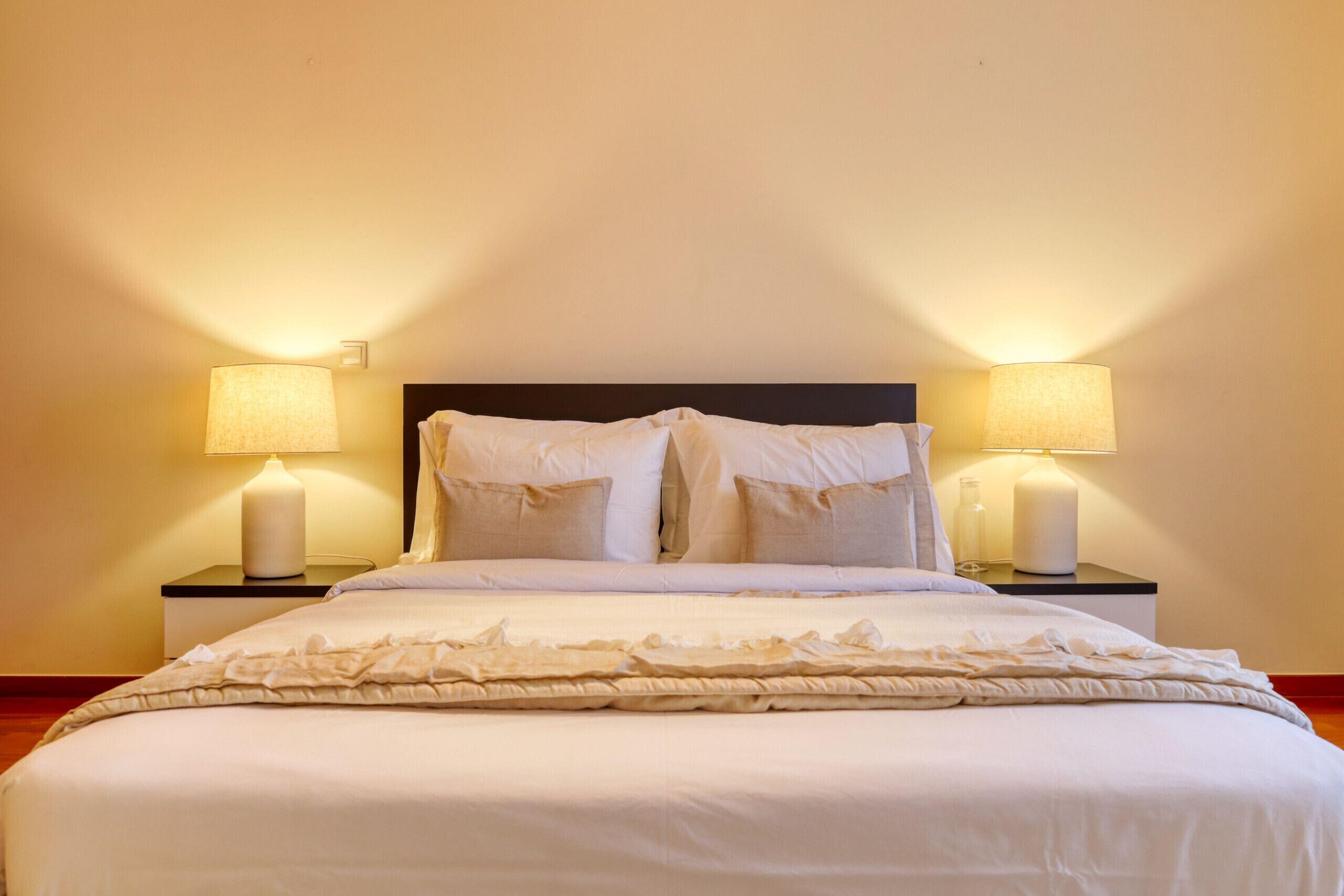 Een net opgemaakt bed met witte lakens en een decoratieve plaid, met aan weerszijden nachtlampjes op bijpassende nachtkastjes.