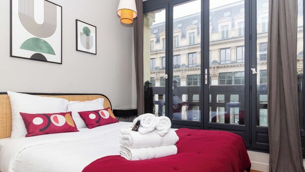 A hotel room with a large bed, a red bedspread, white bedding, and a view of buildings through a large window. - Elegant Apartment - Montorgueil - Le Marais (Paris)