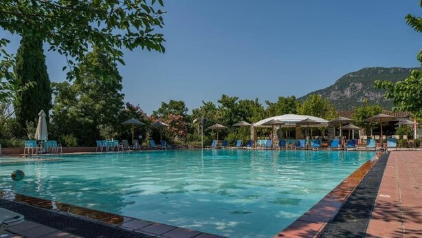 Piscina con sillas reclinables y sombrillas, rodeada de árboles y una montaña al fondo.