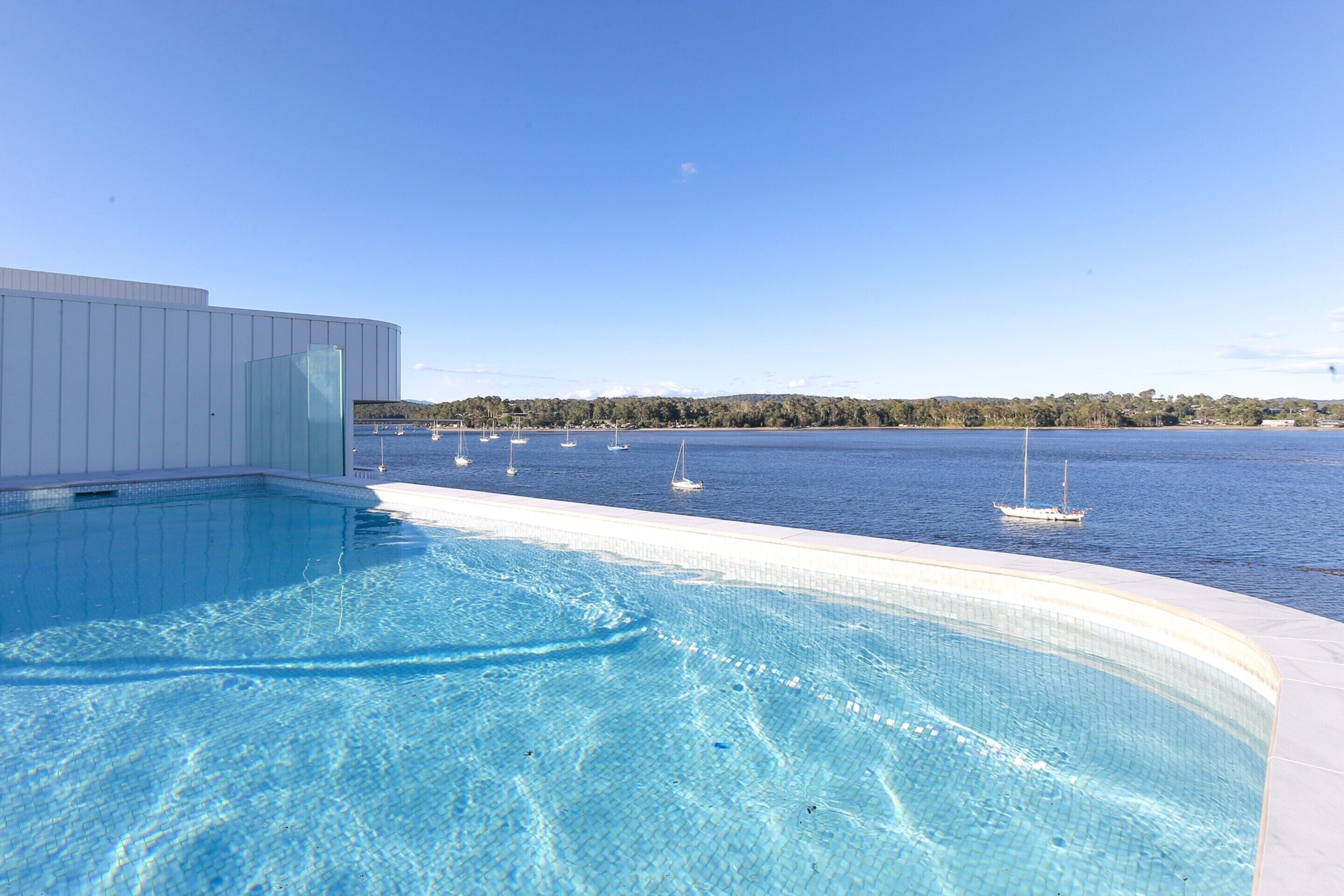 A swimming pool with clear blue water, surrounded by a white pool deck and a white building. In the background, there is a view of a calm sea with several sailboats and a distant shoreline with trees.