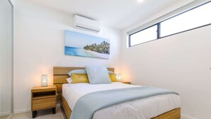 A bedroom with a bed, nightstands, a wall-mounted air conditioner, a window, and a beach scene painting. - Stunning 1-bed Bayside Apartment With Superb Views (Batemans Bay)