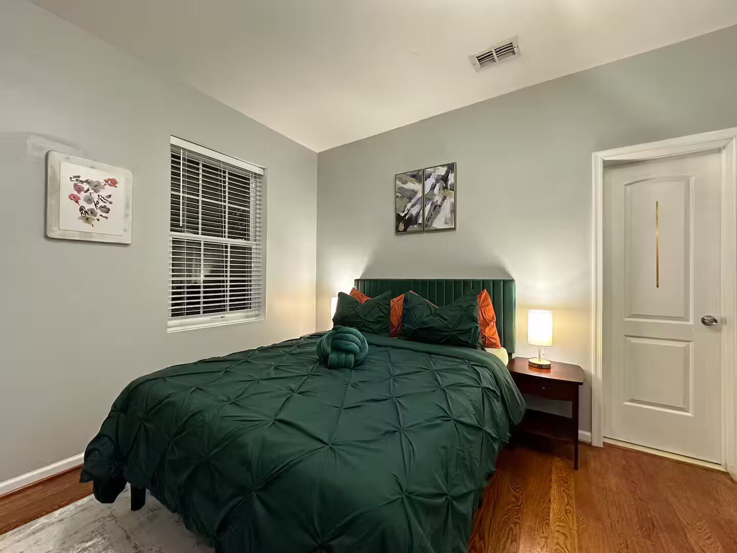 A bedroom with a green bedspread, a window with blinds, a door, and a nightstand with a lamp.