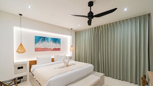 Habitación de hotel moderna con una cama grande, un ventilador de techo, un cuadro y vista al océano tras unas cortinas.