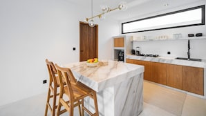 Una cocina moderna con una isla de mármol, sillas de madera, un frutero y una lámpara colgante.