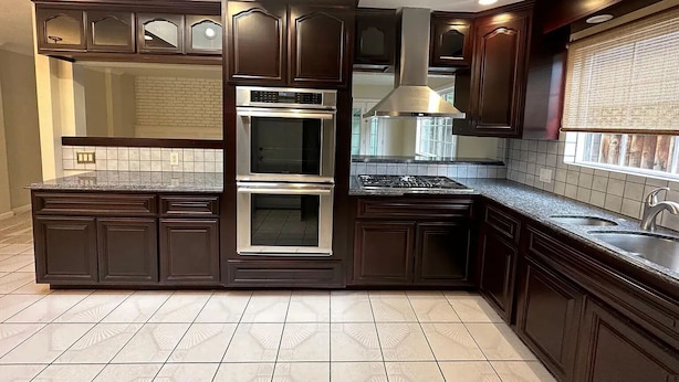 Une cuisine avec des meubles de couleur bois foncé, un four double, une cuisinière en acier inoxydable et une fenêtre avec des stores.