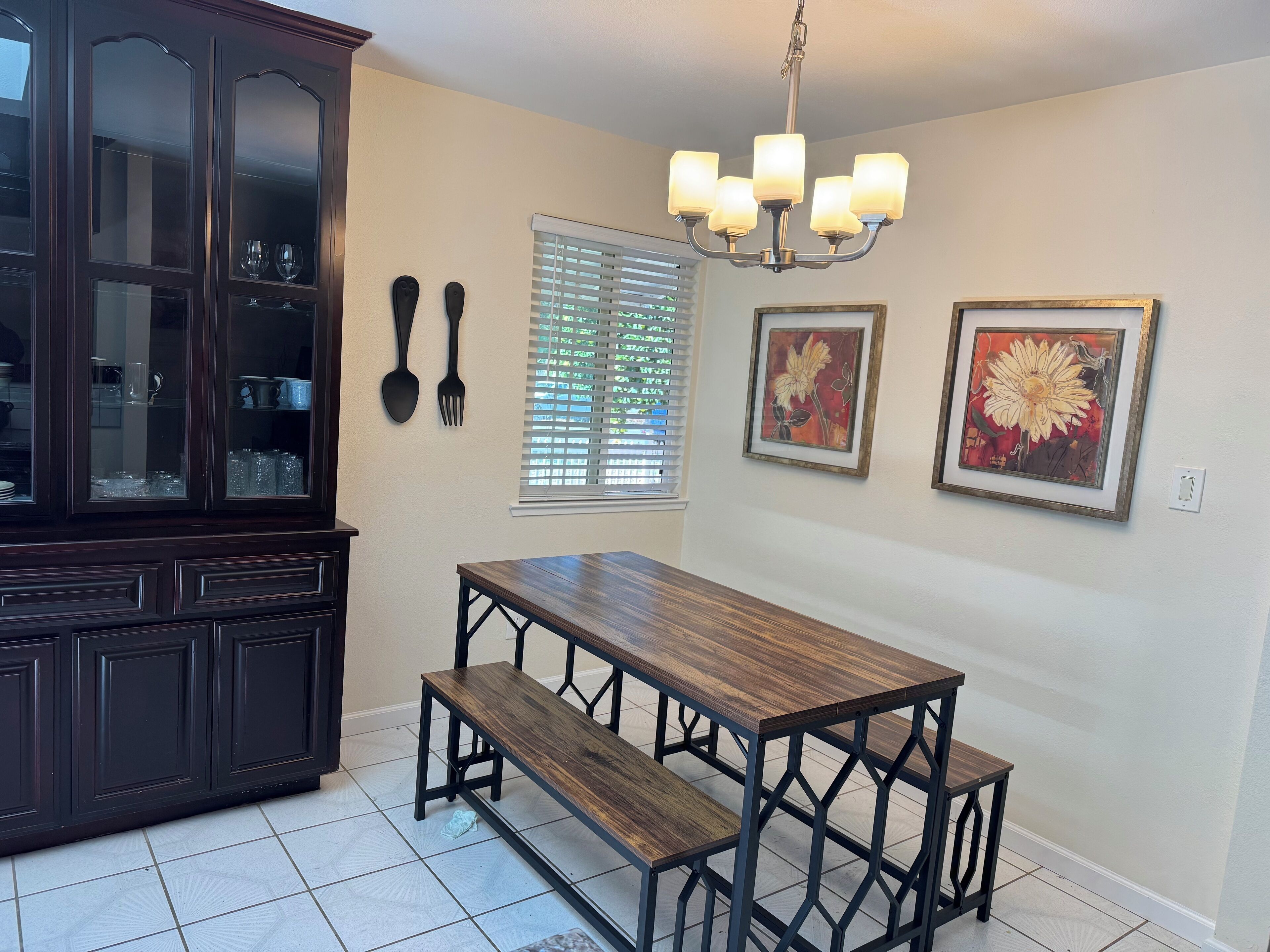 A dining area with a dark wooden table and benches, a glass-fronted cabinet, a chandelier, and two framed floral paintings on the wall.
