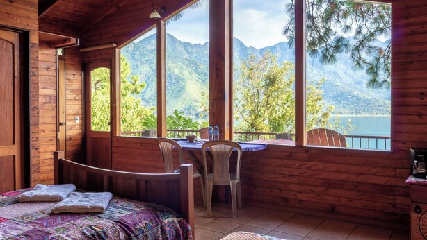 Habitación de cabaña de madera con cama, una mesa con sillas y grandes ventanas que ofrecen vista a la montaña.